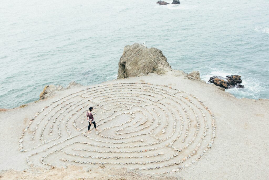 Person geht durch ein aus Steinen gelegtes Labyrinth an einer felsigen Küste am Meer.
